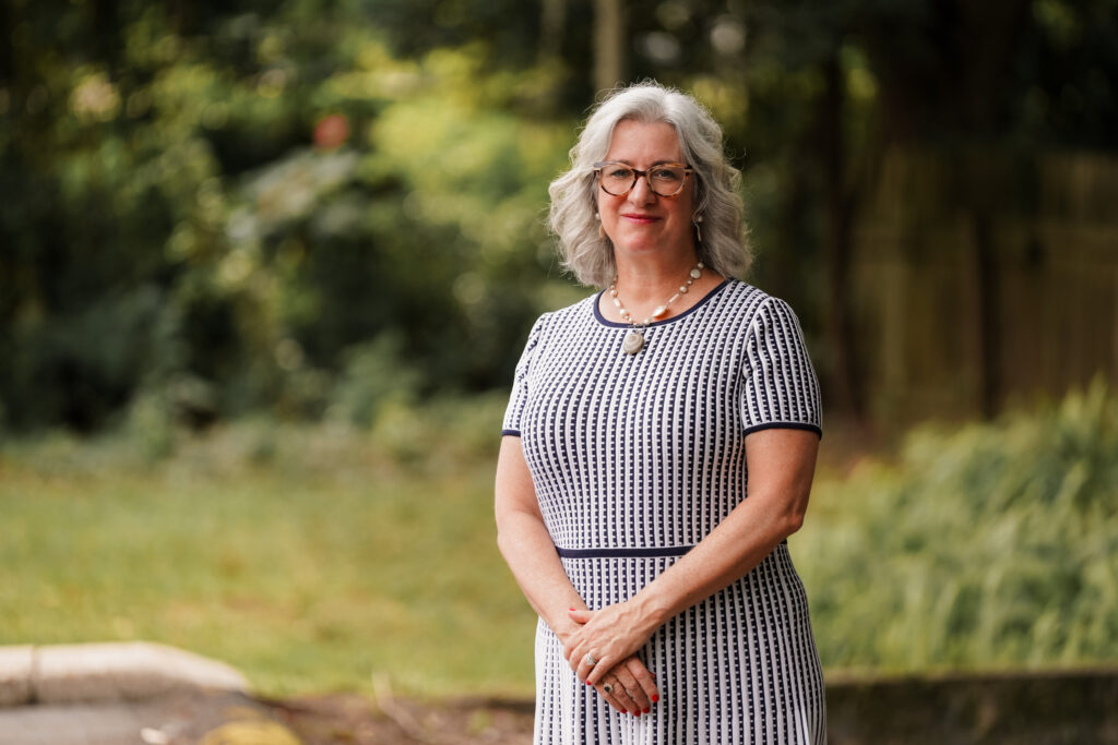 Managing Partner, Shannon Novey, standing outside, smiling, with greenery behind her